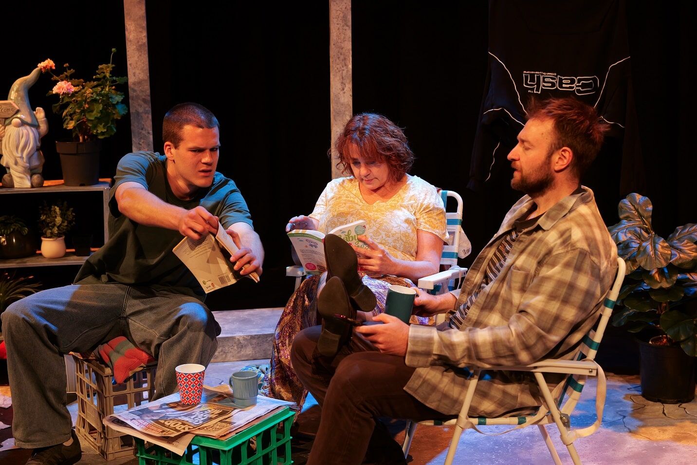 Three adults seated in a cozy, cluttered set: man on the left hands over papers, woman in the middle reads a magazine, man on the right chats; cups and newspapers on a crate-table between them with plants and a gnome statue in the background.